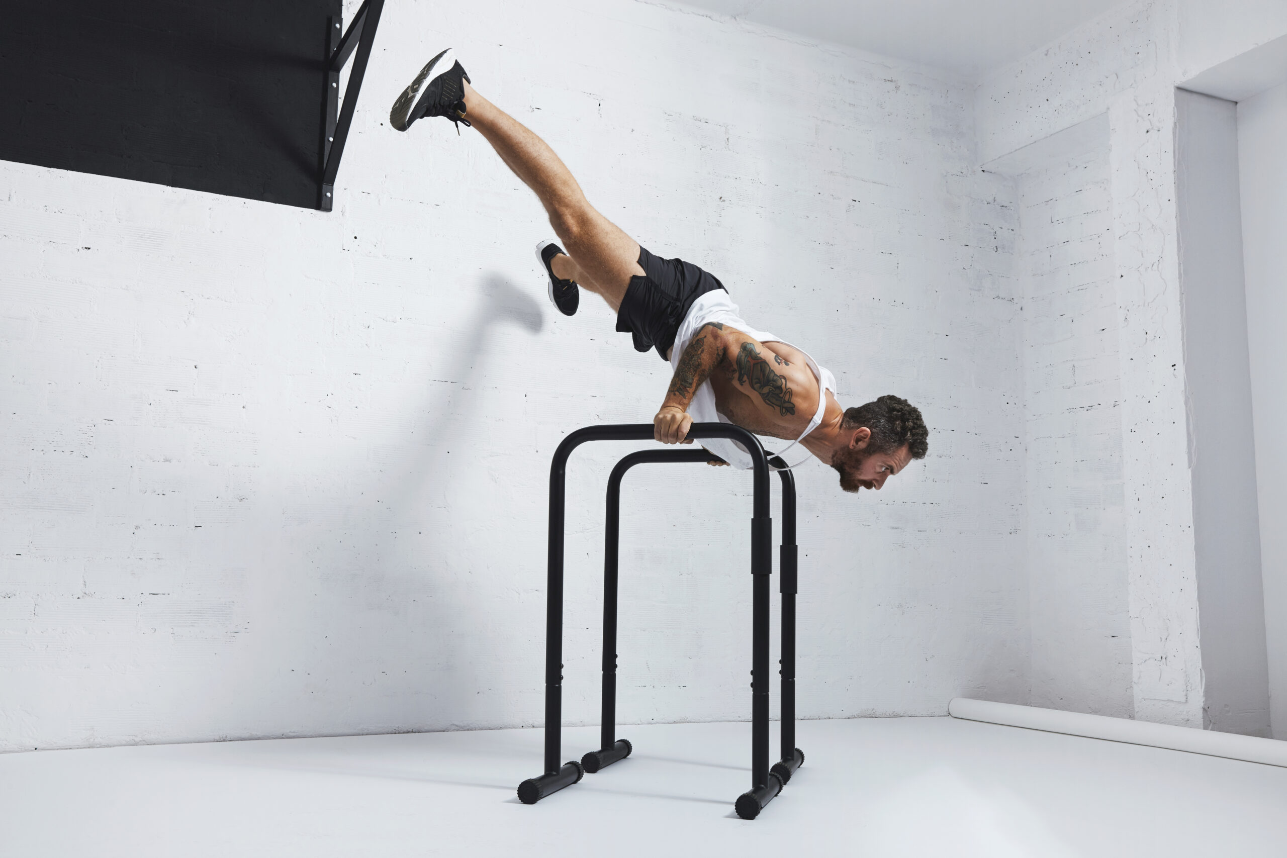 Strong tattooed in white unlabeled tank t-shirt male athlete shows calisthenic moves Extended legs planche push ups on parallel bars